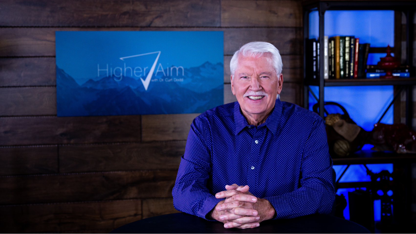 Dr. Curt Dodd in blue shirt sitting in front of a wooden wall with 'Higher Aim' logo.